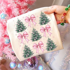 May include: A cream-colored tote bag featuring a repeating pattern of pink bows, green Christmas trees with pink ornaments, and small white snowflakes. The bag is held by a person with a bracelet of red, green, and gold bells.
