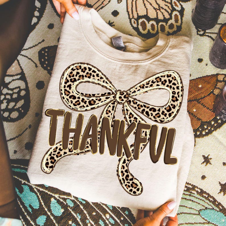 May include: A beige sweatshirt with a large bow design featuring a leopard print pattern. The word "THANKFUL" is printed in a brown, bold font below the bow. The sweatshirt is folded and held by hands.