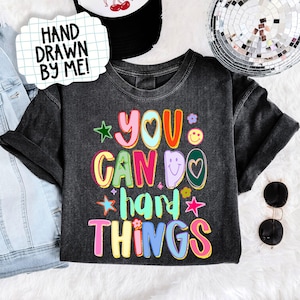 May include: A dark gray t-shirt with the phrase "YOU CAN DO HARD THINGS" in colorful, bubble letters. The shirt has a crew neck and short sleeves. A white sign with the text "HAND DRAWN BY ME!" is in the upper left corner.