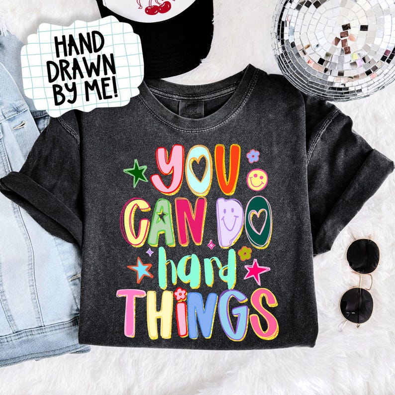 May include: A dark gray t-shirt with the phrase "YOU CAN DO HARD THINGS" in colorful, bubble letters. The shirt has a crew neck and short sleeves. A white sign with the text "HAND DRAWN BY ME!" is in the upper left corner.