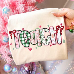 May include: A cream-colored t-shirt with the word "TEACH" in a festive, Christmas-themed design. The letters are filled with patterns like green plaid, gingerbread men, and snowflakes, and are outlined in pink with red bows.