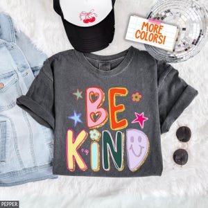 Pode incluir: Uma t-shirt cinza-carvão com o texto BE KIND em letras coloridas e brilhantes. O design inclui corações cor-de-rosa, um K amarelo, um I azul-claro, um N verde-escuro e uma cara sorridente roxa clara. Um boné de cerejas, um casaco de ganga e óculos de sol estão por perto.