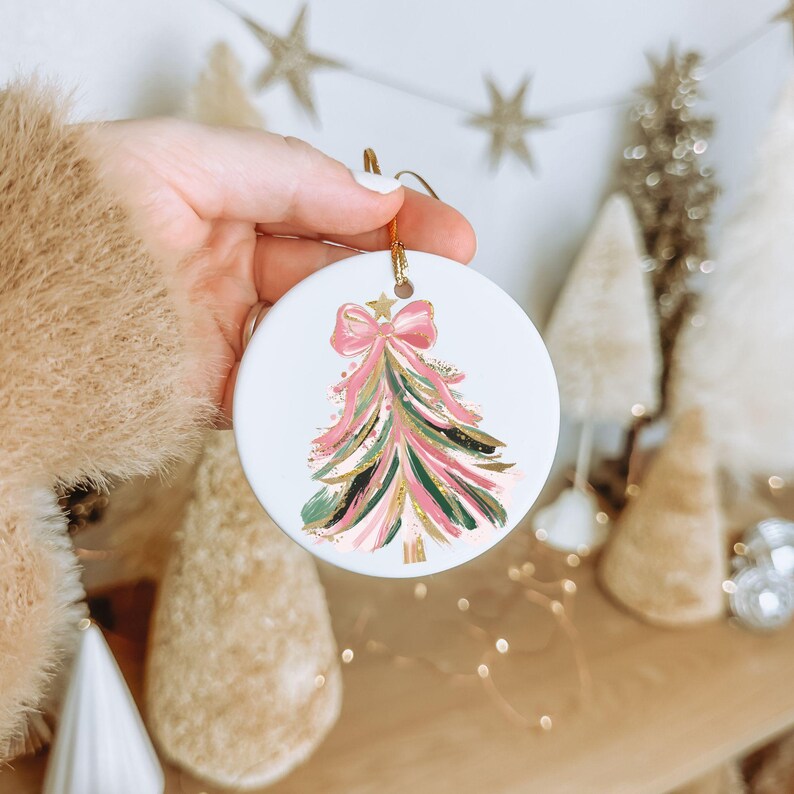 May include: White ceramic Christmas ornament with a hand-painted Christmas tree design. The tree features pink, green, and gold brushstrokes, topped with a pink bow and gold star. A gold string is attached for hanging.