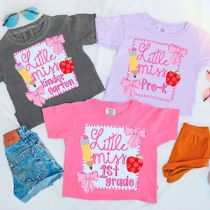 Op de afbeelding: Drie shirts met korte mouwen in grijs, lavendel en roze, elk met een schoolthema-ontwerp. Het ontwerp bevat een potlood, een appel en de woorden "Little Miss" gevolgd door "Kindergarten", "Pre-K" en "1st Grade".
