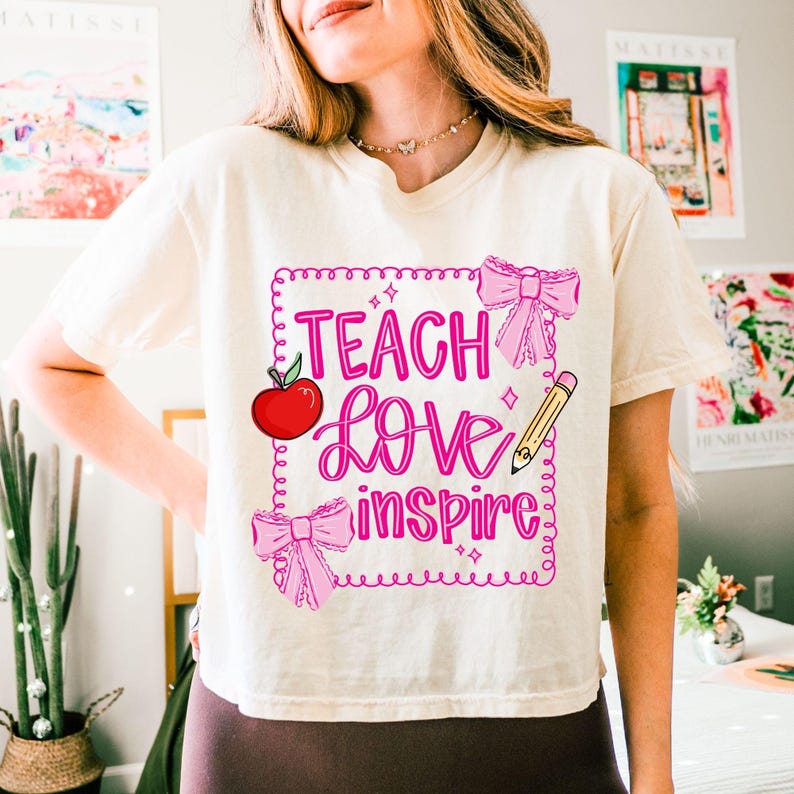 Puede incluir: Camiseta color crema con las palabras "Teach Love Inspire" en escritura rosa dentro de un cuadrado con contorno rosa. El dise&ntilde;o incluye una manzana roja, lazos rosas y un l&aacute;piz amarillo. La camiseta es de estilo casual y de manga corta.