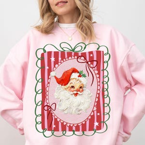 Op de afbeelding: Een lichtroze sweatshirt met een vintage kerstman-ontwerp. De kerstman-afbeelding is omlijst met een rode en groene rand, en de kerstman draagt een rode hoed en heeft een witte baard. Dit is een feestelijk vakantiekledingstuk.