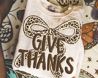 May include: A beige sweatshirt with a brown leopard print bow and the words "GIVE THANKS" in brown block letters. The sweatshirt is folded and held by hands, with a patterned background.