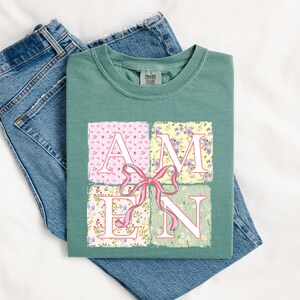 May include: A teal green t-shirt with a floral print design that spells out the word "AMEN". The letters are white with pink and green floral accents. A pink bow is in the center of the design.