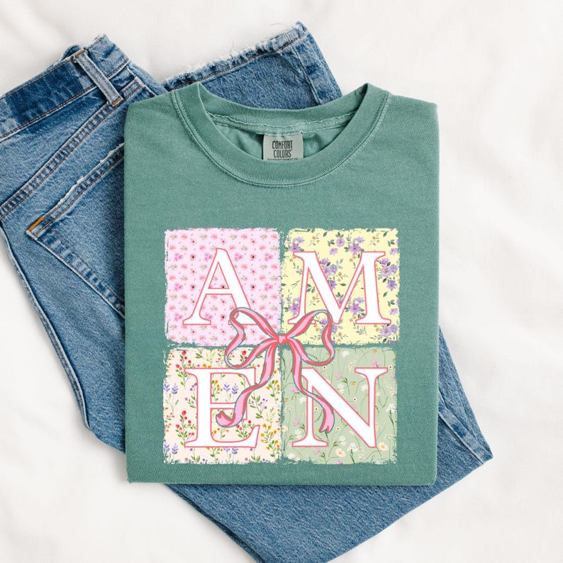 May include: A teal green t-shirt with a floral print design that spells out the word "AMEN". The letters are white with pink and green floral accents. A pink bow is in the center of the design.