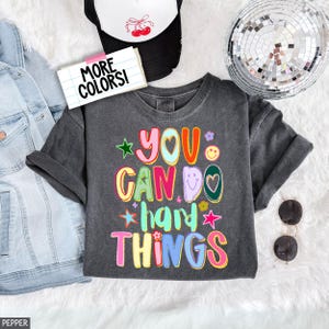 Può includere: T-shirt grigio scuro con la frase "YOU CAN DO hard THINGS" in lettere colorate e giocose. Il design include stelle, fiori e un cuore. Un cappellino da camionista bianco e nero e occhiali da sole sono anche nell'immagine.