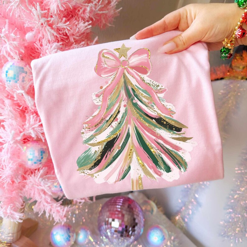 May include: A pink t-shirt featuring a Christmas tree design. The tree is decorated with pink, green, and gold brushstrokes, topped with a gold star and a pink bow. The shirt is held up against a pink Christmas tree.