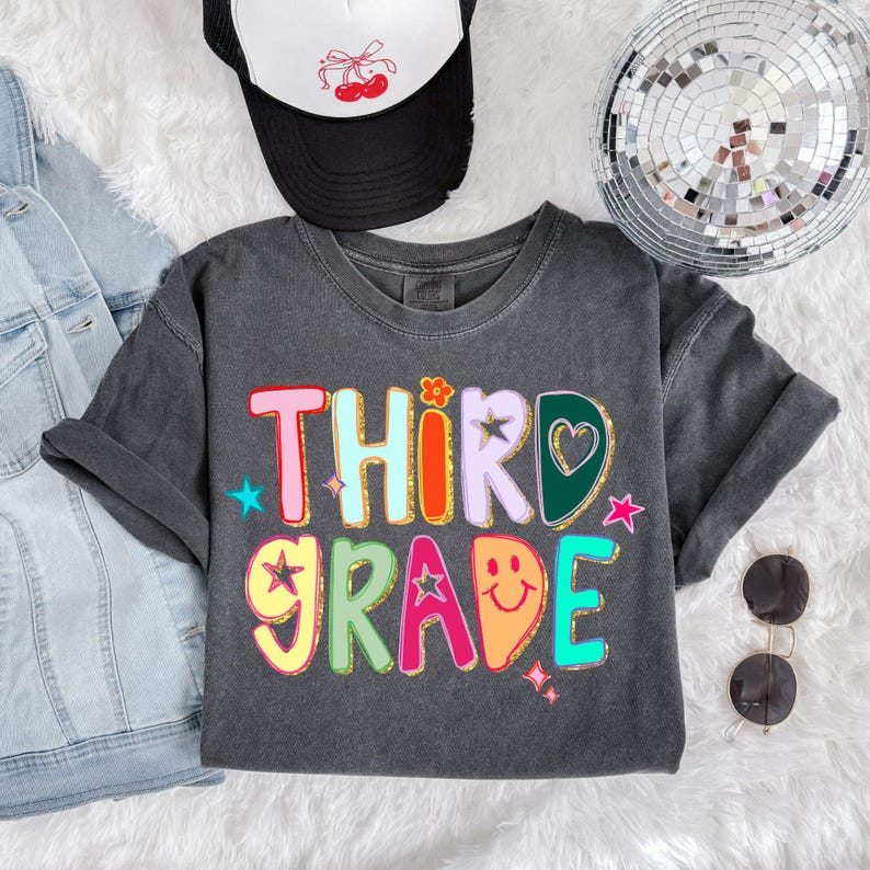 May include: A dark grey t-shirt with the words "THIRD GRADE" in colourful, glittery letters. The design includes stars, a heart, and a smiley face. A black and white trucker hat with a cherry design is visible.