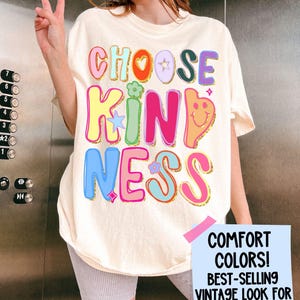 May include: Ivory-colored t-shirt with the words "CHOOSE KINDNESS" in colorful, bubble letters. The shirt has a vintage look and features a smiley face and flower graphics. The text "COMFORT COLORS! BEST-SELLING VINTAGE LOOK FOR SCHOOL DAYS" is also visible.