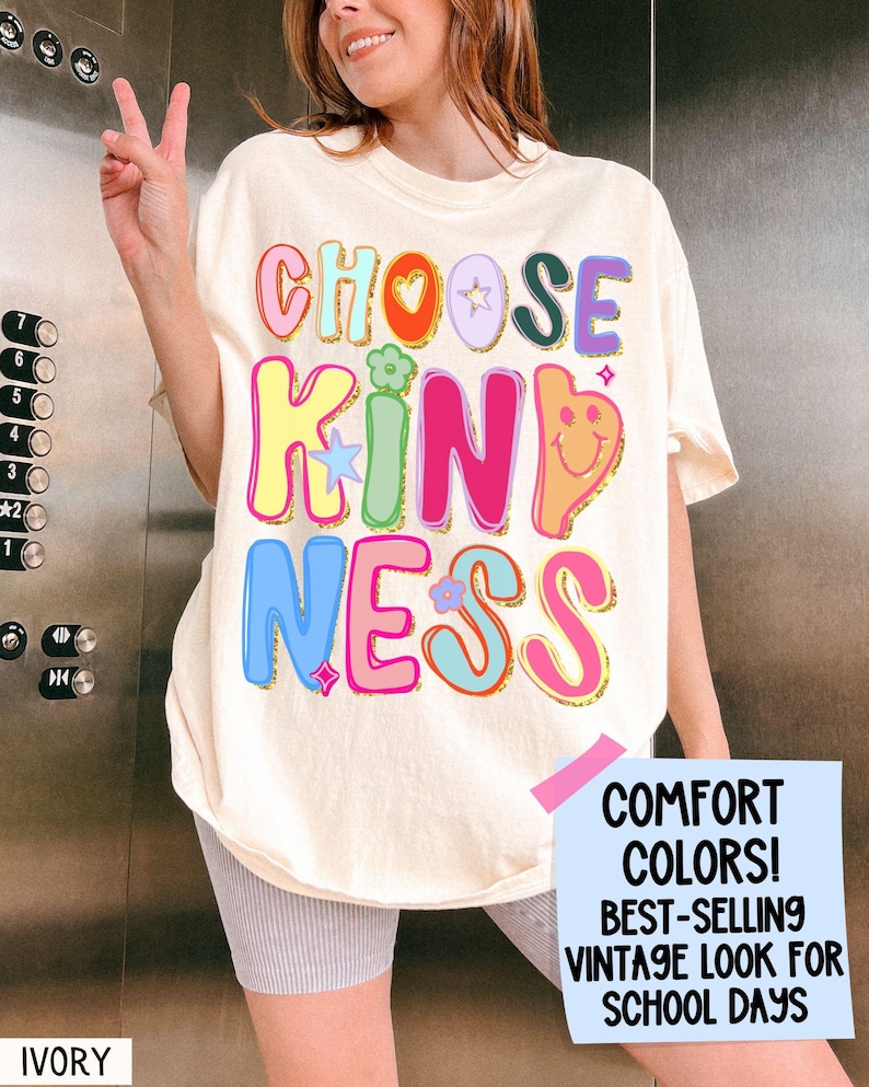 May include: Ivory-colored t-shirt with the words "CHOOSE KINDNESS" in colorful, bubble letters. The shirt has a vintage look and features a smiley face and flower graphics. The text "COMFORT COLORS! BEST-SELLING VINTAGE LOOK FOR SCHOOL DAYS" is also visible.
