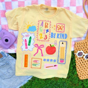K&ouml;nnte beinhalten: Ein hellgelbes Kurzarm-T-Shirt mit einem farbenfrohen Grafikdruck von Illustrationen zum Thema Schule, darunter ein Apfel, ein Bleistift, ein Lineal, Buntstifte, B&uuml;cher, eine Sonne, Wolken, Schleifen und der Text "BE KIND" neben Buchstaben und Zahlen.