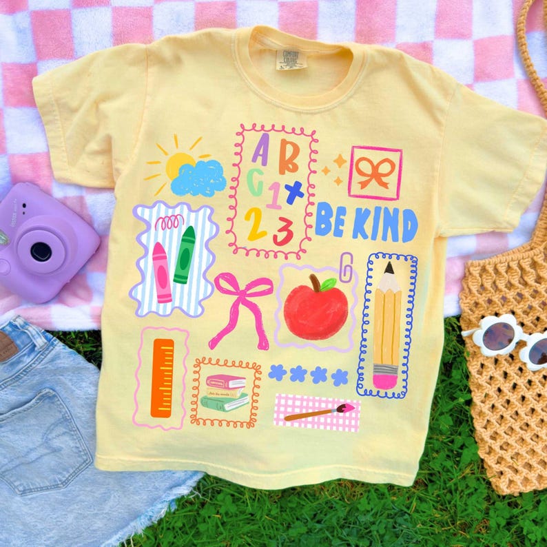 K&ouml;nnte beinhalten: Ein hellgelbes Kurzarm-T-Shirt mit einem farbenfrohen Grafikdruck von Illustrationen zum Thema Schule, darunter ein Apfel, ein Bleistift, ein Lineal, Buntstifte, B&uuml;cher, eine Sonne, Wolken, Schleifen und der Text "BE KIND" neben Buchstaben und Zahlen.