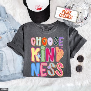 May include: A dark gray t-shirt with the words "CHOOSE KINDNESS" in colorful, bubble letters. The design includes a heart, star, flower, and smiley face. A black and white trucker hat and sunglasses are also visible.