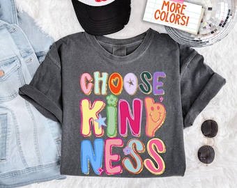 Könnte beinhalten: Ein dunkelgraues T-Shirt mit dem Schriftzug "CHOOSE KINDNESS" in bunten Buchstaben. Das Design beinhaltet ein Herz, einen Stern, eine Blume und ein Smiley-Gesicht. Eine schwarz-weiße Trucker-Cap und Sonnenbrille sind ebenfalls zu sehen.