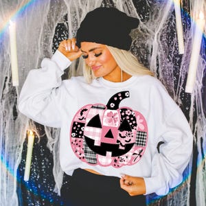May include: White sweatshirt featuring a patchwork pumpkin design in pink, black, and white. The pumpkin has a black stem and face, with floral, plaid, and bat patterns. A black beanie is worn.