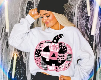 May include: White sweatshirt featuring a patchwork pumpkin design in pink, black, and white. The pumpkin has a black stem and face, with floral, plaid, and bat patterns. A black beanie is worn.