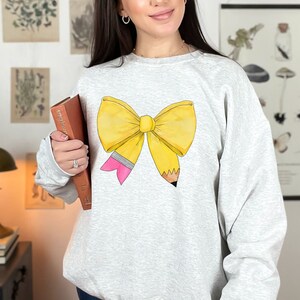 May include: A gray sweatshirt with a yellow pencil bow design. The bow is tied with a pink ribbon and the pencil is sharpened.