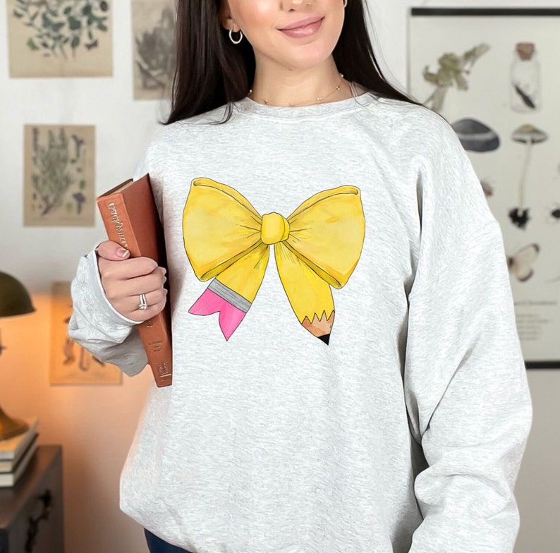 May include: A gray sweatshirt with a yellow pencil bow design. The bow is tied with a pink ribbon and the pencil is sharpened.