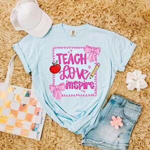 Puede incluir: Camiseta azul claro con las palabras "Teach Love Inspire" en rosa, junto con una manzana roja, lazos rosas y un l&aacute;piz amarillo. La camiseta se combina con pantalones cortos de mezclilla, una gorra de b&eacute;isbol blanca y un bolso de mano.