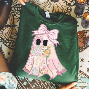 Puede incluir: Sudadera verde oscuro con un aplique de fantasma. El diseño del fantasma está hecho de tela de retazos con estampados florales y cuadros rosas, y un lazo rosa. El fantasma tiene ojos ovalados negros. Una prenda acogedora y única.