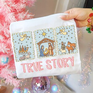 May include: White t-shirt featuring a nativity scene illustration with a light blue background and gold trim. The design includes angels, the three wise men, and the text "TRUE STORY" in pink.