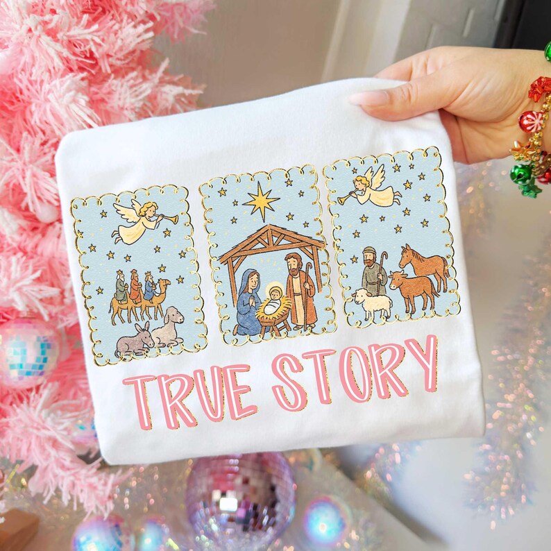 May include: White t-shirt featuring a nativity scene illustration with a light blue background and gold trim. The design includes angels, the three wise men, and the text "TRUE STORY" in pink.