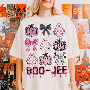 Może przedstawiać: Kremowa koszulka z grafiką o tematyce Halloween. Projekt zawiera r&oacute;żowe i czarne dynie, kokardy i duchy z wzorami w szachownicę. Tekst "BOO-JEE" jest wydrukowany kroplami, idealny na upiorny sezon.