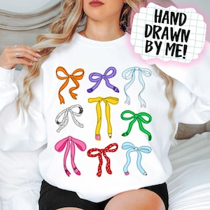 May include: White sweatshirt with a pattern of colourful bows in orange, purple, and pink. The text "HAND DRAWN BY ME!" is in a pink bubble in the upper right corner.