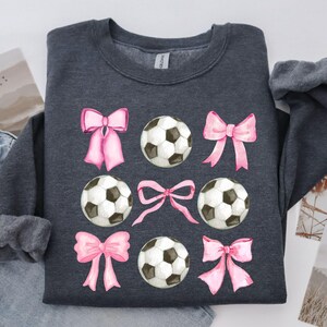 May include: Dark gray sweatshirt featuring a pattern of soccer balls and pink bows. The soccer balls are black and white, and the bows are a soft pink. The sweatshirt is a casual style.