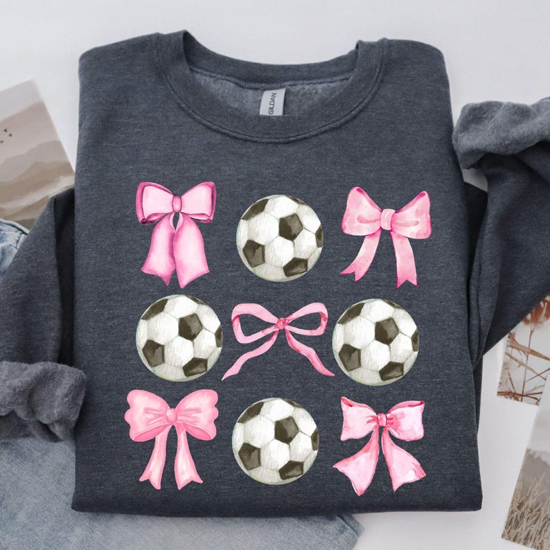 May include: Dark gray sweatshirt featuring a pattern of soccer balls and pink bows. The soccer balls are black and white, and the bows are a soft pink. The sweatshirt is a casual style.