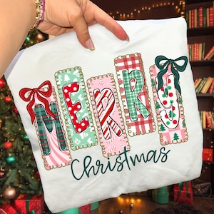 Può includere: Felpa bianca a tema natalizio con la parola "MERRY" scritta in lettere colorate e decorate, ciascuna con un bordo decorativo. La parola "Christmas" è scritta sotto in un carattere corsivo. Un fiocco rosso adorna la lettera "M".