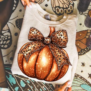 Puede incluir: Una sudadera beige con un diseño de calabaza naranja brillante y un lazo con estampado de leopardo. La calabaza tiene un efecto brillante y el lazo está adornado con pedrería. La sudadera está doblada y sostenida por manos.