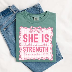 May include: A teal green t-shirt with a pink and white gingham design. The design features a bow and the text "She is clothed with strength Proverbs 31:25".