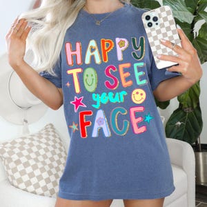 May include: A blue t-shirt with the words "HAPPY TO SEE your FACE" in colorful, bubble letters. The design includes a flower, stars, and smiley faces. The shirt is a casual, comfortable style.