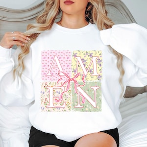 May include: White sweatshirt with a floral print design spelling out the word "AMEN" in block letters. A pink ribbon is tied in the center of the design.
