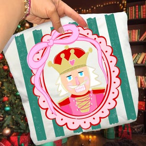 May include: A white cushion with vertical teal stripes and a pink bow. The cushion features a pink and red framed illustration of a nutcracker wearing a gold crown. The nutcracker has a white wig and a pink uniform.