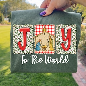 May include: A dark green sweatshirt with the word "JOY" in red letters, framed with holly and berries. The letter "O" features a nativity scene with Mary, Joseph, and baby Jesus. The text "To The World" is written below in white script.