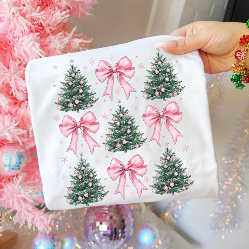 May include: White t-shirt featuring a repeating pattern of green Christmas trees adorned with pink ornaments and bows. Small pink snowflakes are scattered around the trees and bows. The design is festive and whimsical.