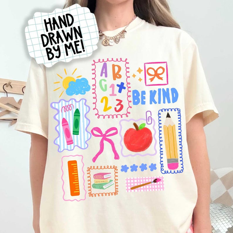 K&ouml;nnte beinhalten: Ein cremefarbenes T-Shirt mit einem bunten Grafikdruck von schulbezogenen Illustrationen, darunter eine Sonne, Buchstaben, Zahlen, Buntstifte, ein roter Apfel, ein Bleistift, ein Lineal, B&uuml;cher und ein Pinsel, mit dem Text "BE KIND" und "HAND DRAWN BY ME!"