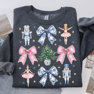 May include: Dark gray sweatshirt featuring a festive design with pink bows, blue and white bows, nutcrackers, ballerinas, a Christmas tree, snowflakes, and gold stars. The design is centered on the front of the sweatshirt.
