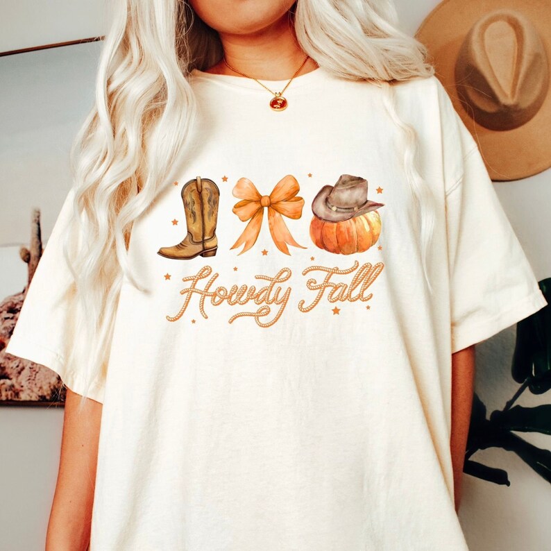 May include: Cream-colored t-shirt featuring a watercolor design with a cowboy boot, bow, and pumpkin with a cowboy hat. The text "Howdy Fall" is written in a rope-like font. The shirt is a relaxed fit.