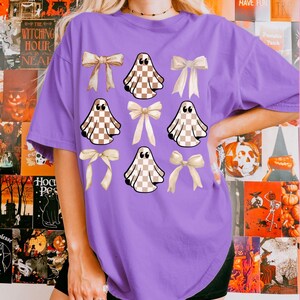 May include: A purple t-shirt featuring a pattern of tan and white checkered ghosts and cream-colored bows. The shirt has a relaxed fit and short sleeves. The background includes Halloween-themed posters with text like "The Witching Hour" and "Girls Just Wanna Have Fun".
