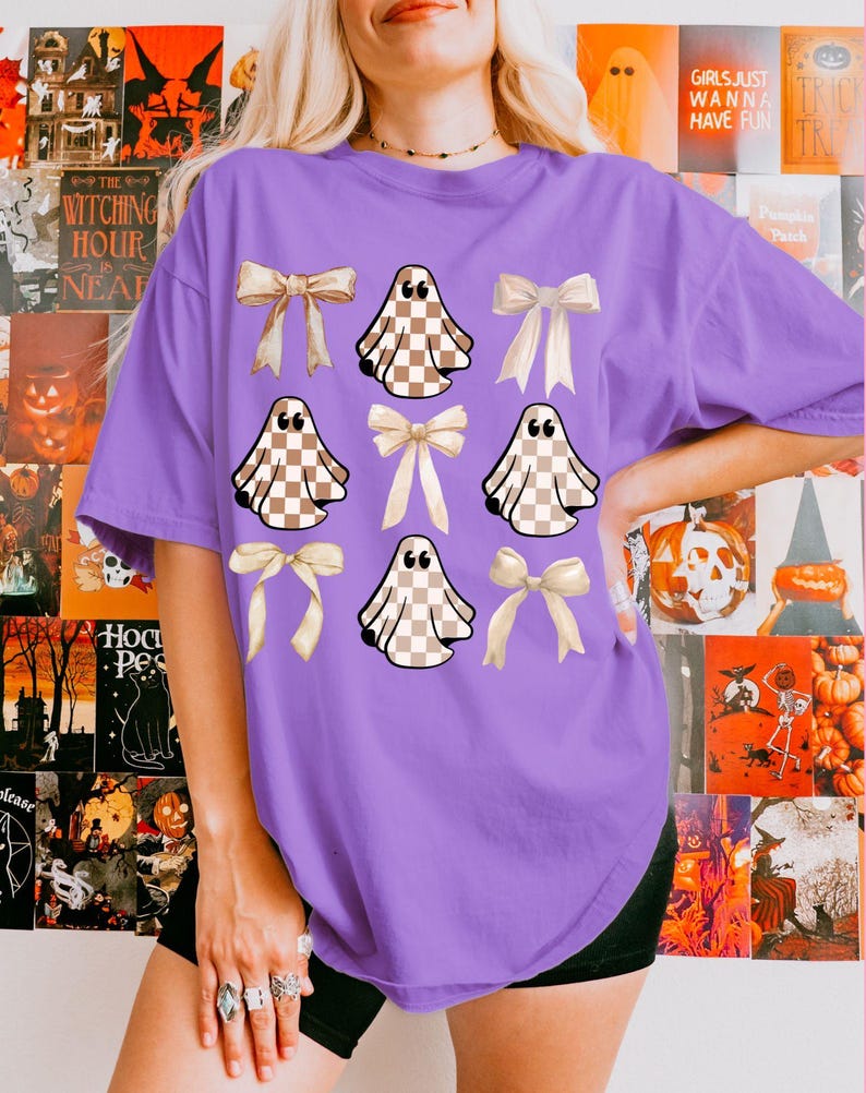 May include: A purple t-shirt featuring a pattern of tan and white checkered ghosts and cream-colored bows. The shirt has a relaxed fit and short sleeves. The background includes Halloween-themed posters with text like "The Witching Hour" and "Girls Just Wanna Have Fun".