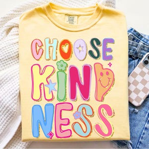 May include: A light yellow t-shirt with the words "CHOOSE KINDNESS" in colorful, bubble letters. The design includes hearts, stars, and a smiling face. A phone with a checkered case rests on blue jeans.