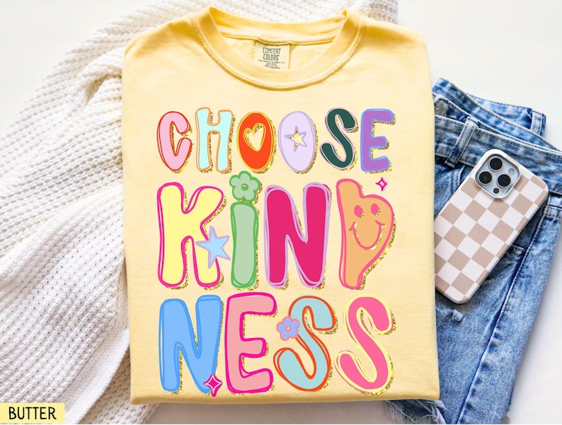 May include: A light yellow t-shirt with the words "CHOOSE KINDNESS" in colorful, bubble letters. The design includes hearts, stars, and a smiling face. A phone with a checkered case rests on blue jeans.