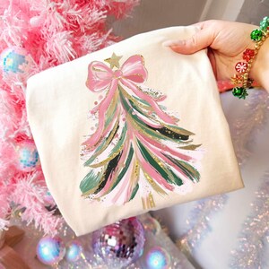 May include: A cream-colored tote bag featuring a Christmas tree design. The tree is composed of pink, green, and gold brushstrokes, topped with a pink bow and gold star. The bag is held in a hand.